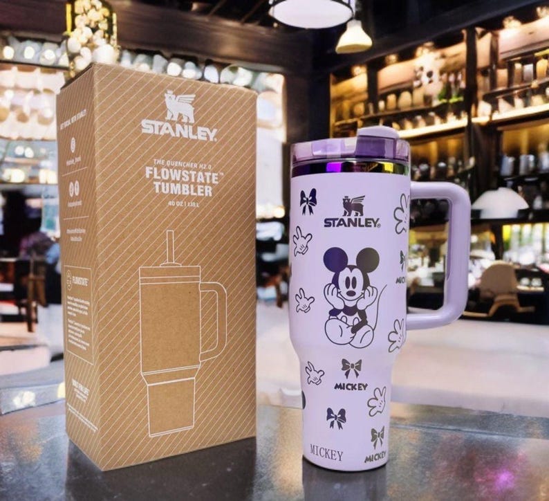 May include: A light purple Stanley Flowstate tumbler with a Mickey Mouse graphic, featuring a handle, a lid, and a straw. The tumbler is displayed next to its brown cardboard packaging, which is printed with the Stanley logo and product name.