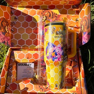 May include: A yellow Stanley tumbler featuring a honeycomb pattern and illustrations of Winnie the Pooh, Piglet, Eeyore, and Tigger. The tumbler includes a yellow handle, a matching lid, and a clear straw, all presented in a matching honeycomb box.