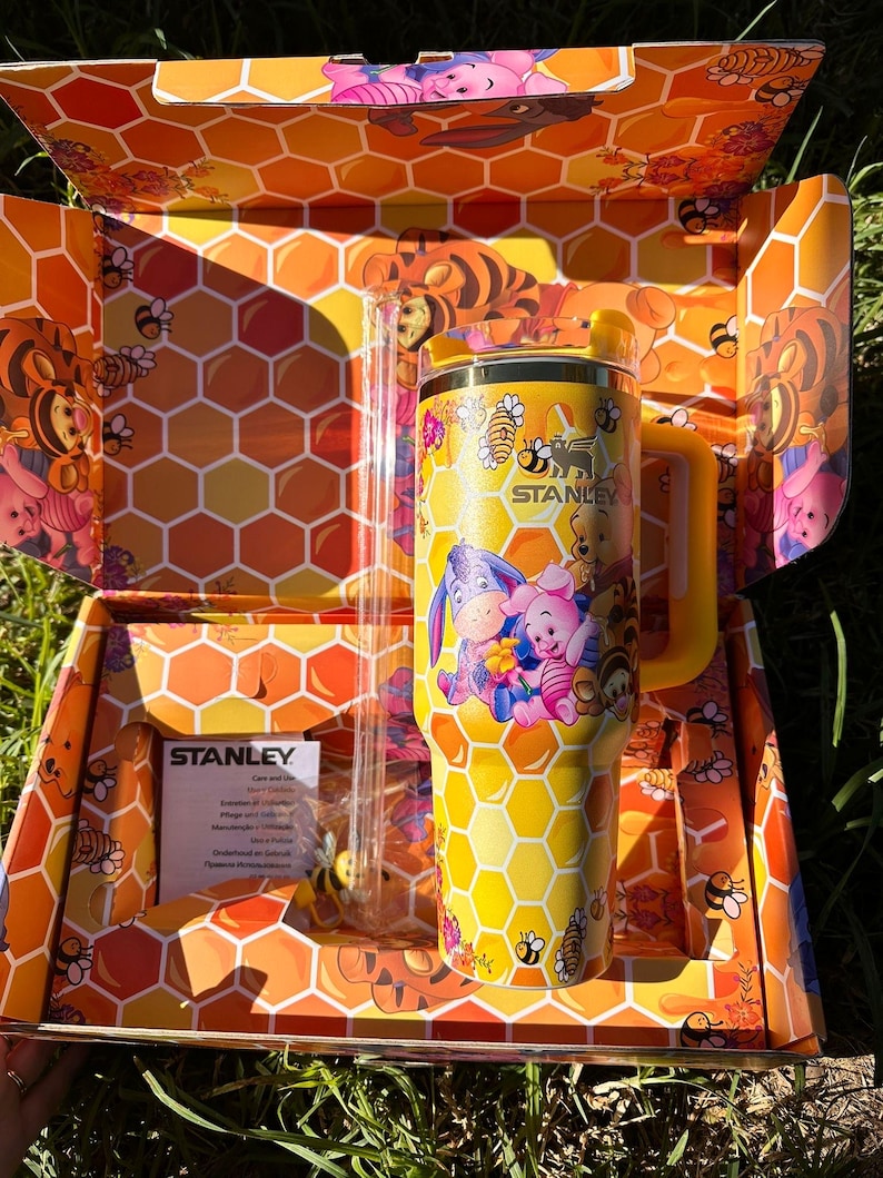 May include: A yellow Stanley tumbler featuring a honeycomb pattern and illustrations of Winnie the Pooh, Piglet, Eeyore, and Tigger. The tumbler includes a yellow handle, a matching lid, and a clear straw, all presented in a matching honeycomb box.