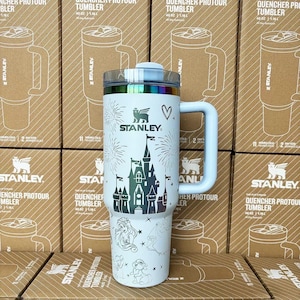 May include: A white Stanley Quencher ProTour tumbler with a 40 oz capacity, featuring a dark graphic of a castle, fireworks, a heart, and various character illustrations. The tumbler has a large side handle, a clear lid, and a metallic rim.