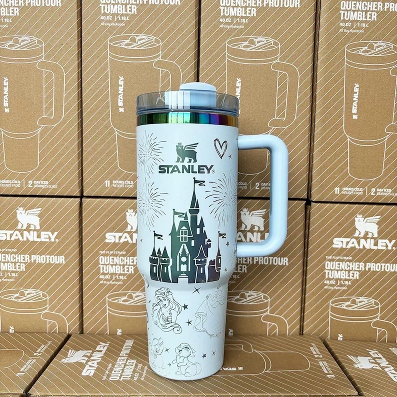 May include: A white Stanley Quencher ProTour tumbler with a 40 oz capacity, featuring a dark graphic of a castle, fireworks, a heart, and various character illustrations. The tumbler has a large side handle, a clear lid, and a metallic rim.