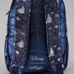 May include: A dark blue backpack featuring a metallic silver pattern of Cinderella, glass slippers, and pumpkins. The shoulder straps are padded and decorated with the same silver print. The Disney and Smiggle logos are printed in white at the base.