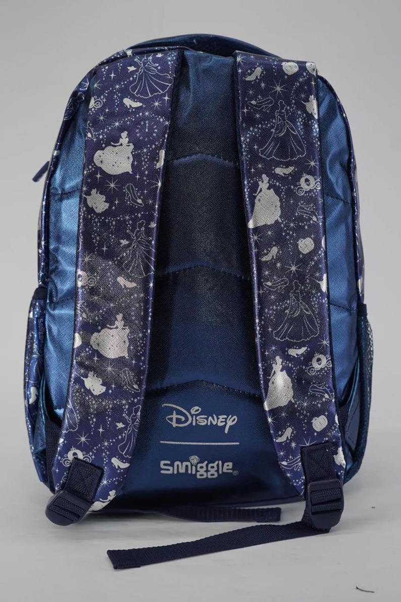 May include: A dark blue backpack featuring a metallic silver pattern of Cinderella, glass slippers, and pumpkins. The shoulder straps are padded and decorated with the same silver print. The Disney and Smiggle logos are printed in white at the base.