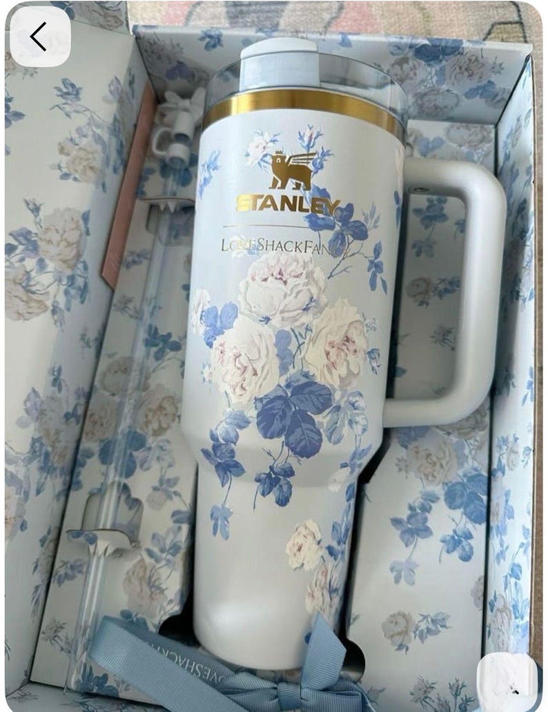May include: A white Stanley tumbler with a blue and white floral pattern, featuring the text Stanley and LoveShackFancy. The tumbler has a gold rim, a clear lid, and a side handle. It is presented in a matching floral box with a clear straw included.