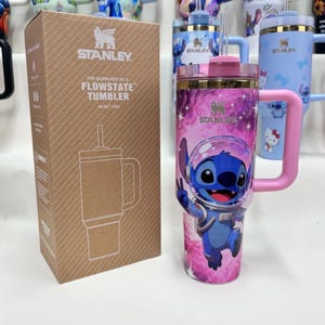 May include: A 40 oz Stanley Quencher H2.0 FlowState tumbler featuring a pink and purple galaxy print with a graphic of Stitch in a space helmet. The tumbler has a pink handle, a clear lid with a straw, and comes in a brown cardboard box.