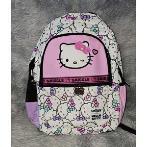 Peut inclure: Un sac &agrave; dos blanc avec un motif Hello Kitty sur l'ensemble, agr&eacute;ment&eacute; de n&oelig;uds roses, violets et bleu sarcelle. La poche avant pr&eacute;sente un grand visage Hello Kitty avec un n&oelig;ud rose paillet&eacute;, une bande noire Smiggle et une &eacute;tiquette Smiggle.