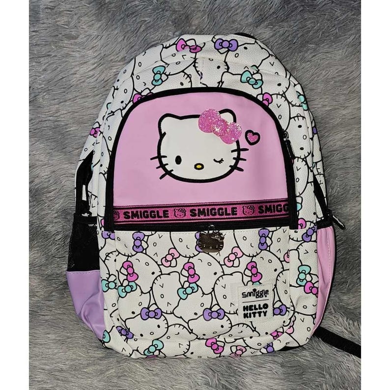 Peut inclure: Un sac &agrave; dos blanc avec un motif Hello Kitty sur l'ensemble, agr&eacute;ment&eacute; de n&oelig;uds roses, violets et bleu sarcelle. La poche avant pr&eacute;sente un grand visage Hello Kitty avec un n&oelig;ud rose paillet&eacute;, une bande noire Smiggle et une &eacute;tiquette Smiggle.