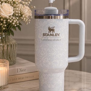K&ouml;nnte beinhalten: Ein hellblauer Stanley LoveShackFancy Becher mit wei&szlig;em Blumen- und Schleifenmuster, einem seitlichen Griff, einem durchsichtigen Deckel und einem Metallstrohhalm. Das Stanley-Logo und der Text LoveShackFancy sind wei&szlig; auf den Becher gedruckt.