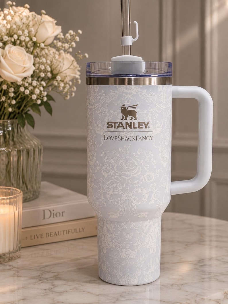 K&ouml;nnte beinhalten: Ein hellblauer Stanley LoveShackFancy Becher mit wei&szlig;em Blumen- und Schleifenmuster, einem seitlichen Griff, einem durchsichtigen Deckel und einem Metallstrohhalm. Das Stanley-Logo und der Text LoveShackFancy sind wei&szlig; auf den Becher gedruckt.