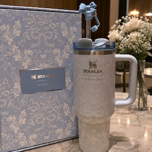 K&ouml;nnte beinhalten: Ein hellblauer und wei&szlig;er Stanley LoveShackFancy Becher mit passender Box. Der Becher hat ein Blumenmuster, einen Griff, einen Deckel und einen Strohhalm mit einer hellblauen Schleife. Die Logos von Stanley und LoveShackFancy sind auf dem Becher und der Box aufgedruckt.