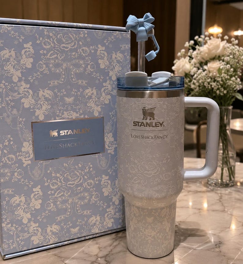 K&ouml;nnte beinhalten: Ein hellblauer und wei&szlig;er Stanley LoveShackFancy Becher mit passender Box. Der Becher hat ein Blumenmuster, einen Griff, einen Deckel und einen Strohhalm mit einer hellblauen Schleife. Die Logos von Stanley und LoveShackFancy sind auf dem Becher und der Box aufgedruckt.