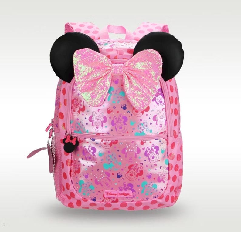 Pode incluir: Uma mochila rosa com uma estampa de Minnie Mouse em toda a superf&iacute;cie e bolinhas nas laterais. A bolsa inclui duas orelhas pretas arredondadas no topo e um grande la&ccedil;o de lantejoulas rosa iridescente centralizado entre as orelhas. O puxador do z&iacute;per tem o formato da cabe&ccedil;a da Minnie.