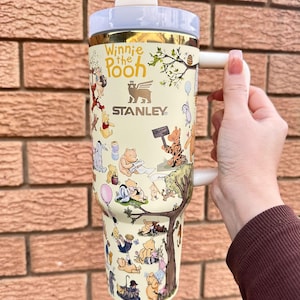 May include: A cream-colored Stanley tumbler featuring Winnie the Pooh characters. The tumbler has a white handle and lid, with gold accents. The design includes illustrations of Pooh, Piglet, Tigger, and Eeyore. The text "Winnie the Pooh" and "Stanley" are visible.