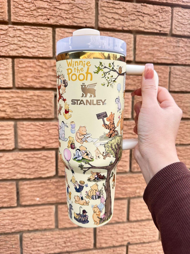 May include: A cream-colored Stanley tumbler featuring Winnie the Pooh characters. The tumbler has a white handle and lid, with gold accents. The design includes illustrations of Pooh, Piglet, Tigger, and Eeyore. The text "Winnie the Pooh" and "Stanley" are visible.