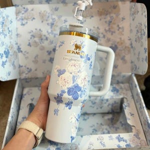May include: A light blue Stanley tumbler with a white floral pattern, a gold rim, and a white handle. The cup features a clear lid with a white straw topped by a small white bow. The text Stanley and LoveShackFancy are printed in gold.