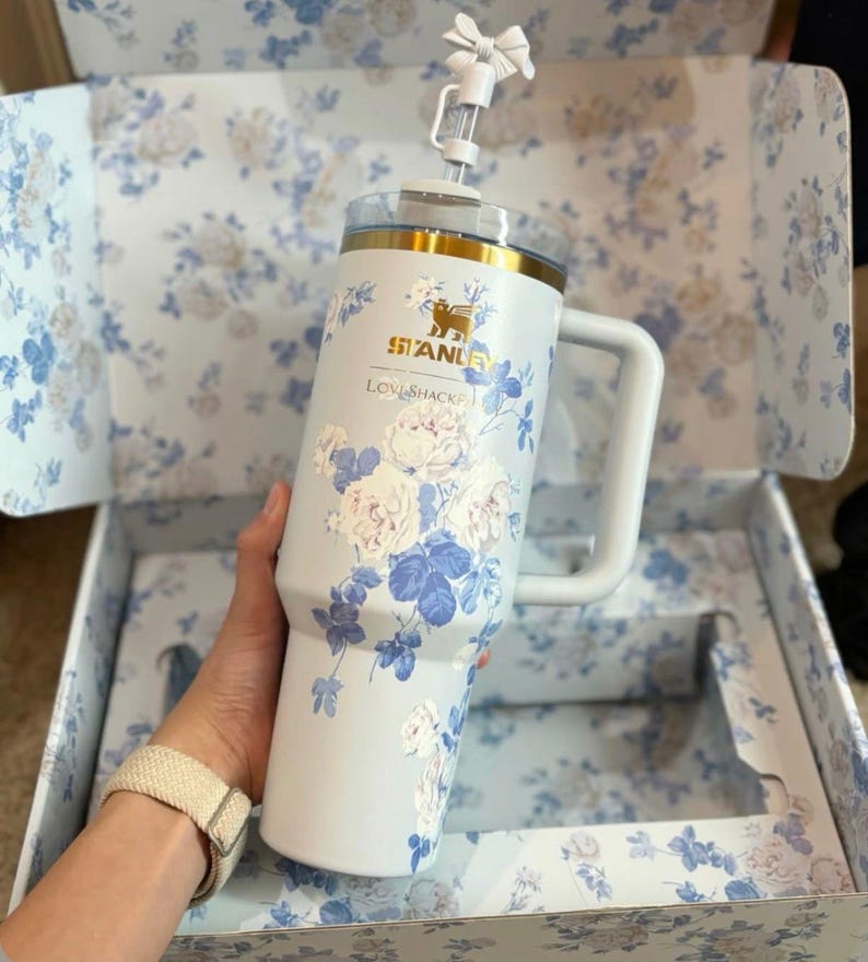 May include: A light blue Stanley tumbler with a white floral pattern, a gold rim, and a white handle. The cup features a clear lid with a white straw topped by a small white bow. The text Stanley and LoveShackFancy are printed in gold.