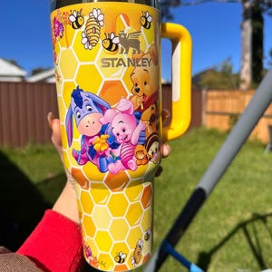May include: A yellow Stanley tumbler featuring a honeycomb pattern and illustrations of Winnie the Pooh, Piglet, Eeyore, and Tigger. The cup includes a yellow handle, a clear lid with a yellow top, and small bee graphics scattered throughout the design.
