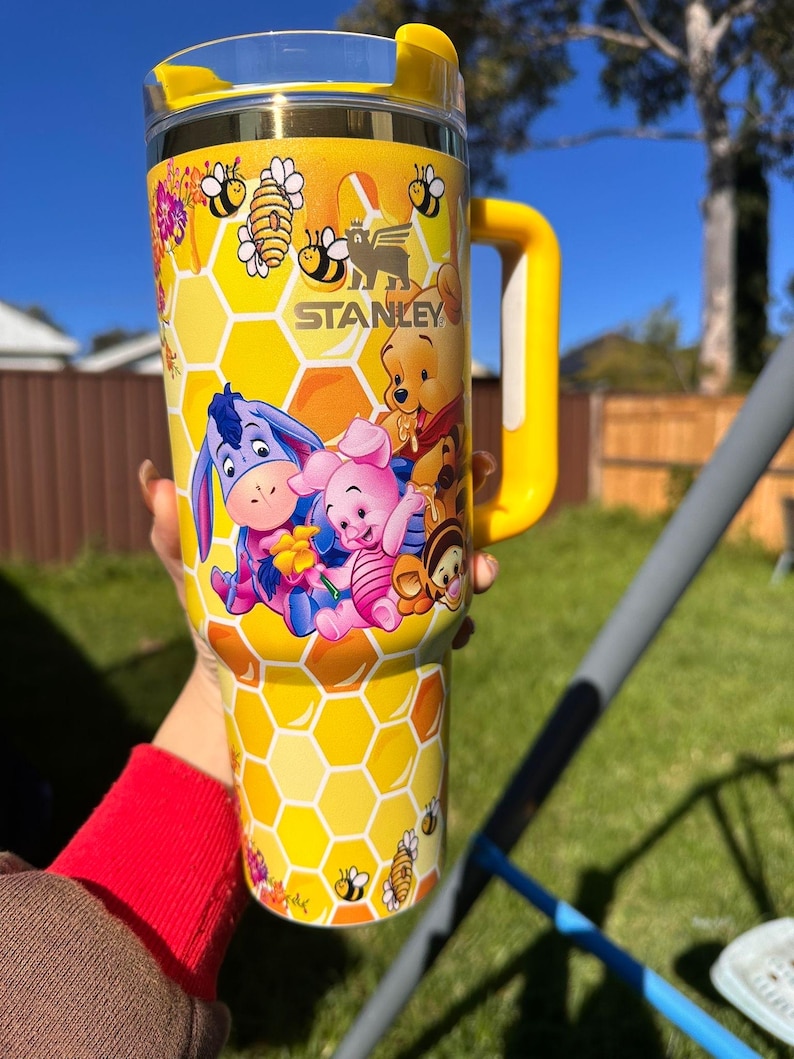May include: A yellow Stanley tumbler featuring a honeycomb pattern and illustrations of Winnie the Pooh, Piglet, Eeyore, and Tigger. The cup includes a yellow handle, a clear lid with a yellow top, and small bee graphics scattered throughout the design.