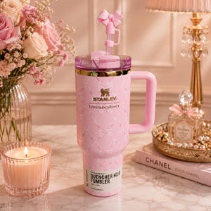 May include: A light pink Stanley Quencher H2.0 tumbler with a 40 oz capacity, featuring a white ribbon pattern, a gold-tone rim, a side handle, and a pink straw topper shaped like a bow. The front displays the Stanley and LoveShackFancy logos.