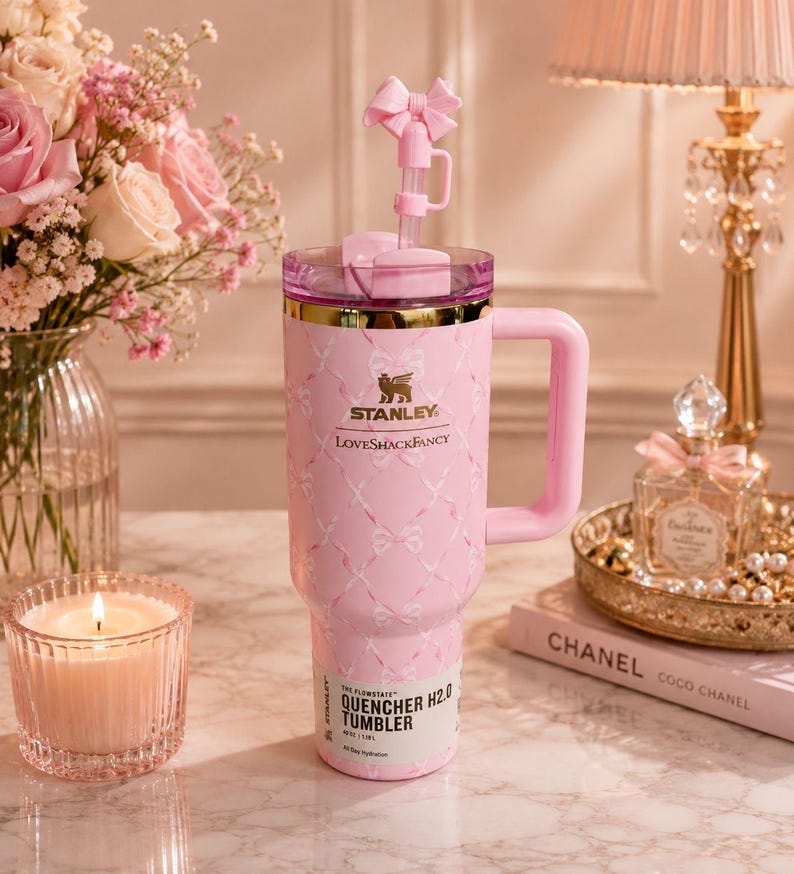May include: A light pink Stanley Quencher H2.0 tumbler with a 40 oz capacity, featuring a white ribbon pattern, a gold-tone rim, a side handle, and a pink straw topper shaped like a bow. The front displays the Stanley and LoveShackFancy logos.