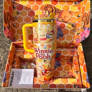 May include: A yellow Stanley tumbler featuring Winnie the Pooh graphics and the text Winnie the Pooh, presented in a matching honeycomb-patterned box with a yellow lid, a side handle, and an included instruction manual in a plastic sleeve.