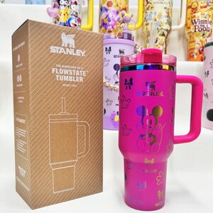 May include: A bright pink Stanley Quencher H2.0 FlowState tumbler with a handle, a clear straw, and a holographic Mickey Mouse graphic. The tumbler is displayed next to its brown cardboard packaging box which features the product name and logo.
