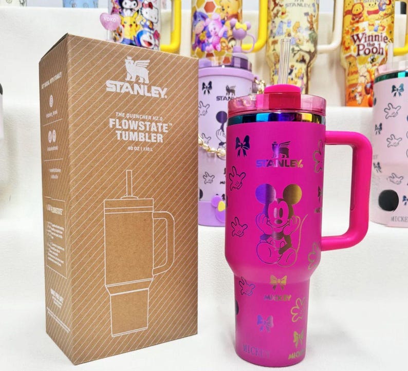 May include: A bright pink Stanley Quencher H2.0 FlowState tumbler with a handle, a clear straw, and a holographic Mickey Mouse graphic. The tumbler is displayed next to its brown cardboard packaging box which features the product name and logo.