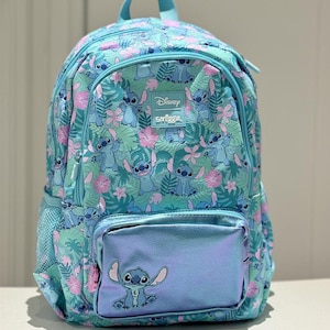 Puede incluir: Una mochila azul claro con un estampado integral del personaje Stitch, flores de hibisco rosas y hojas tropicales verdes. La bolsa incluye un bolsillo frontal con acabado azul met&aacute;lico, un gr&aacute;fico de Stitch y una etiqueta de Disney Smiggle.