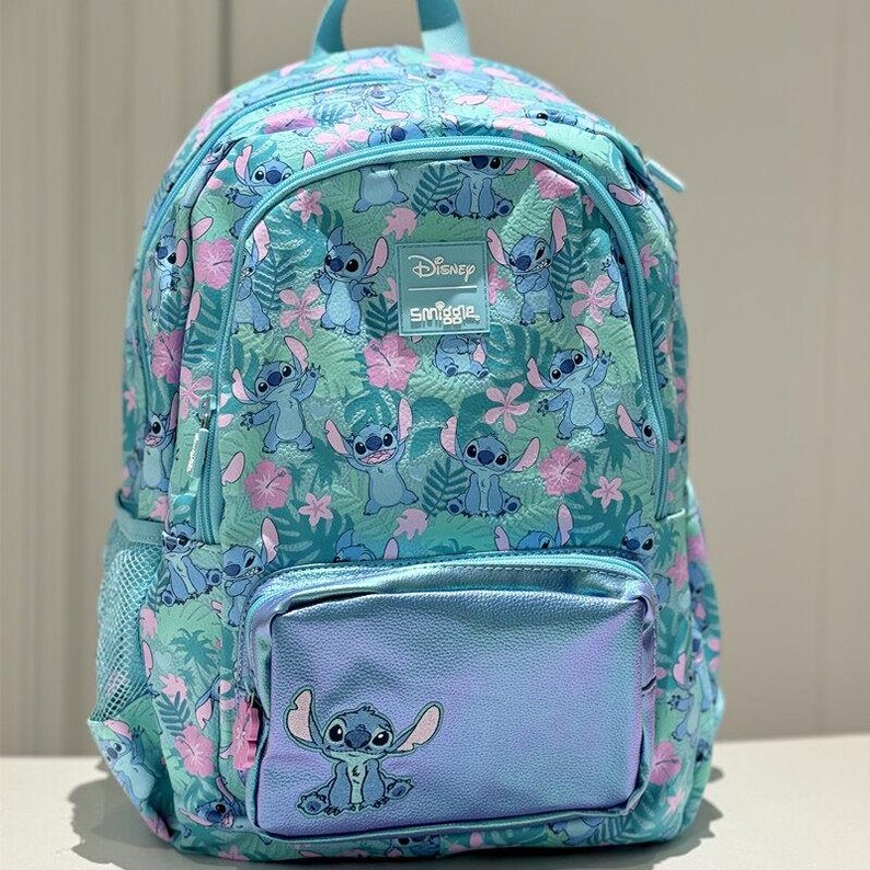 Puede incluir: Una mochila azul claro con un estampado integral del personaje Stitch, flores de hibisco rosas y hojas tropicales verdes. La bolsa incluye un bolsillo frontal con acabado azul met&aacute;lico, un gr&aacute;fico de Stitch y una etiqueta de Disney Smiggle.