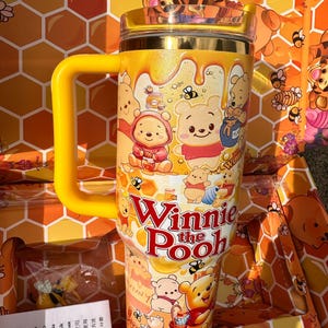May include: A yellow insulated tumbler with a handle featuring a Winnie the Pooh graphic print, honey drip patterns, and bees. The text Winnie the Pooh is printed in red on the front. The tumbler has a clear lid and a yellow handle.