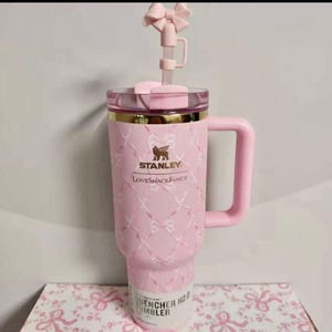 May include: A light pink Stanley LoveShackFancy Quencher tumbler featuring a white ribbon and bow pattern, a gold rim, a matching pink handle, and a pink bow straw topper. The tumbler has the Stanley and LoveShackFancy logos printed in gold.