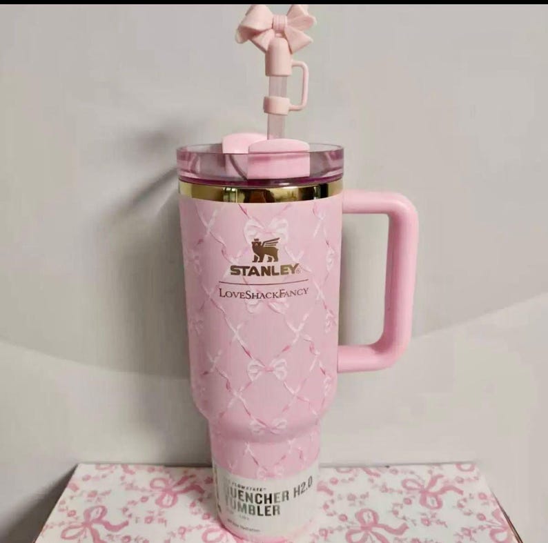 May include: A light pink Stanley LoveShackFancy Quencher tumbler featuring a white ribbon and bow pattern, a gold rim, a matching pink handle, and a pink bow straw topper. The tumbler has the Stanley and LoveShackFancy logos printed in gold.