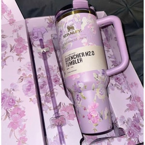 May include: A lavender Stanley Quencher H2.0 tumbler with a floral pattern, featuring a matching handle, lid, and a clear straw with bow accents. The product label reads Stanley, LoveShackFancy, and The Flowstate Quencher H2.0 Tumbler 40 oz | 1.18 L.