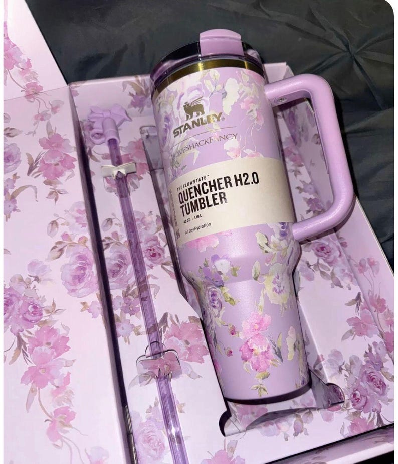 May include: A lavender Stanley Quencher H2.0 tumbler with a floral pattern, featuring a matching handle, lid, and a clear straw with bow accents. The product label reads Stanley, LoveShackFancy, and The Flowstate Quencher H2.0 Tumbler 40 oz | 1.18 L.