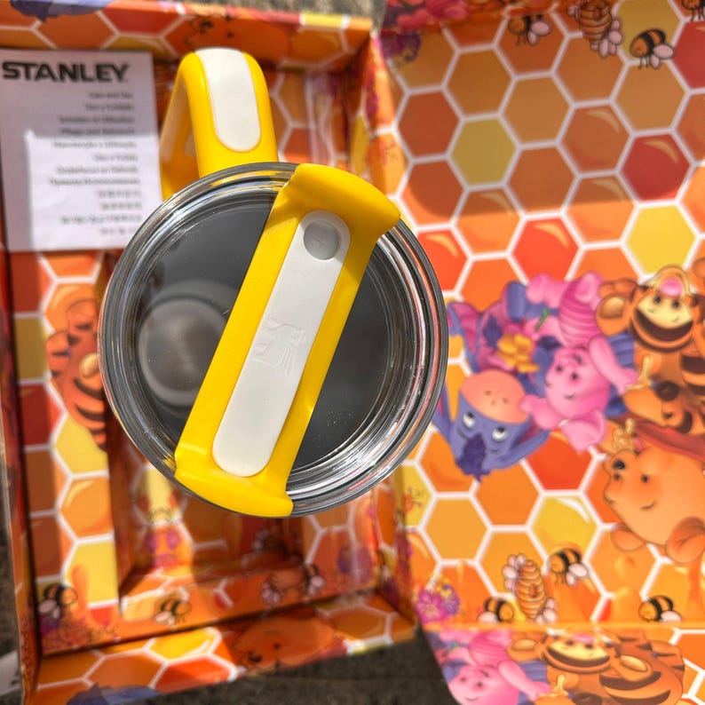 May include: A Stanley brand tumbler lid featuring a bright yellow and white rotating closure mechanism. The lid is circular with a clear plastic rim and is positioned against a box interior decorated with a honeycomb pattern and Winnie the Pooh characters.