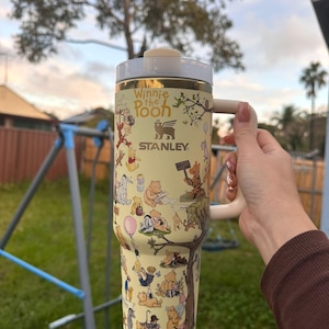 May include: A cream-colored Stanley tumbler featuring Winnie the Pooh characters. The tumbler has a cream-colored handle and lid, with gold accents. The design includes illustrations of Pooh, Piglet, and other characters. The text "Winnie the Pooh" and "Stanley" are visible.