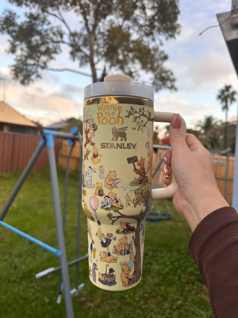 May include: A cream-colored Stanley tumbler featuring Winnie the Pooh characters. The tumbler has a cream-colored handle and lid, with gold accents. The design includes illustrations of Pooh, Piglet, and other characters. The text "Winnie the Pooh" and "Stanley" are visible.