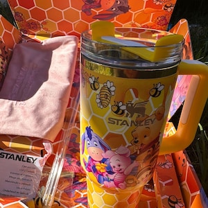 May include: A yellow Stanley tumbler featuring a honeycomb pattern with Winnie the Pooh, Piglet, and Eeyore graphics, a yellow handle, and a clear lid with a yellow rotating cover, presented in a matching honeycomb-patterned cardboard box.