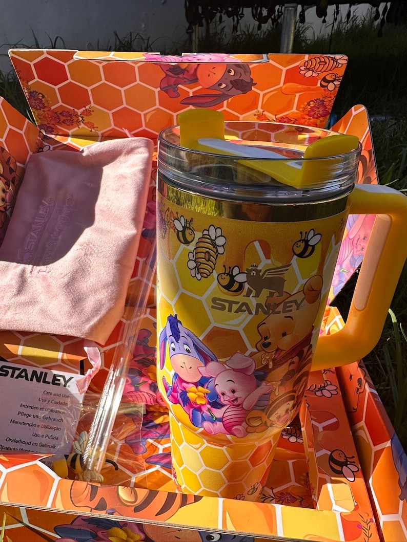 May include: A yellow Stanley tumbler featuring a honeycomb pattern with Winnie the Pooh, Piglet, and Eeyore graphics, a yellow handle, and a clear lid with a yellow rotating cover, presented in a matching honeycomb-patterned cardboard box.