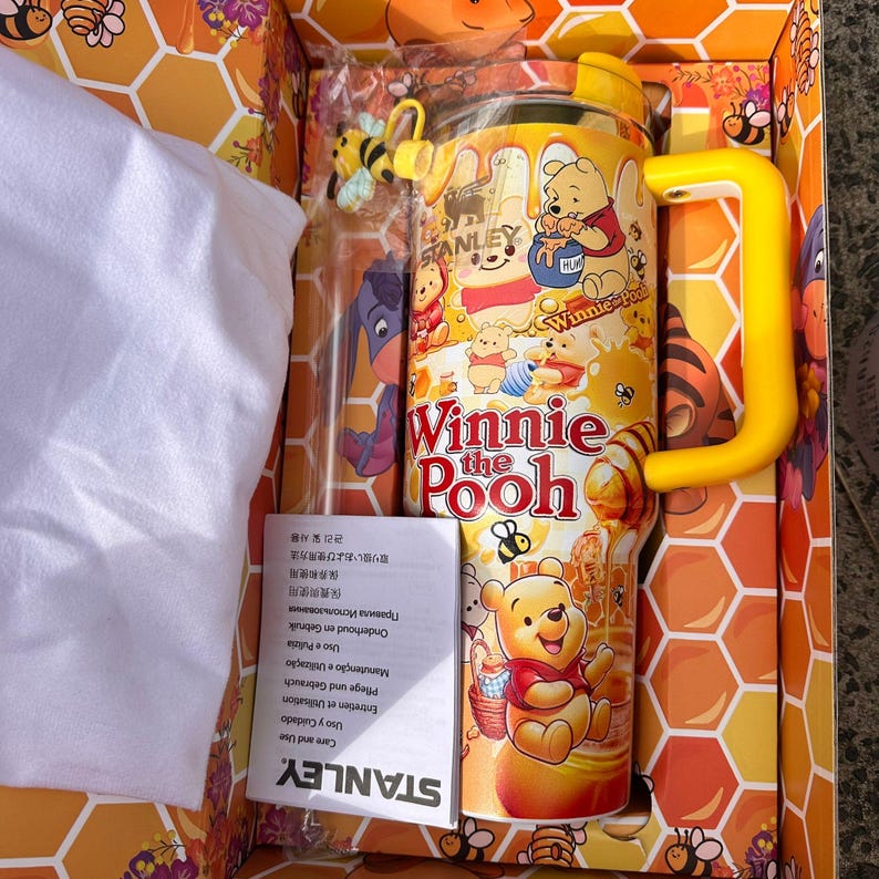 May include: A yellow Stanley tumbler featuring Winnie the Pooh graphics, honey drips, and bees. The cup includes a yellow handle, a lid, and a small bee charm. It is presented in a box with a honeycomb pattern and includes a white instruction manual.