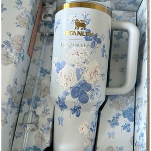 May include: A white Stanley tumbler with a gold rim and a blue and white floral pattern. The cup features the text Stanley and LoveShackFancy. It includes a matching clear straw with a white floral topper, all packaged in a floral box.