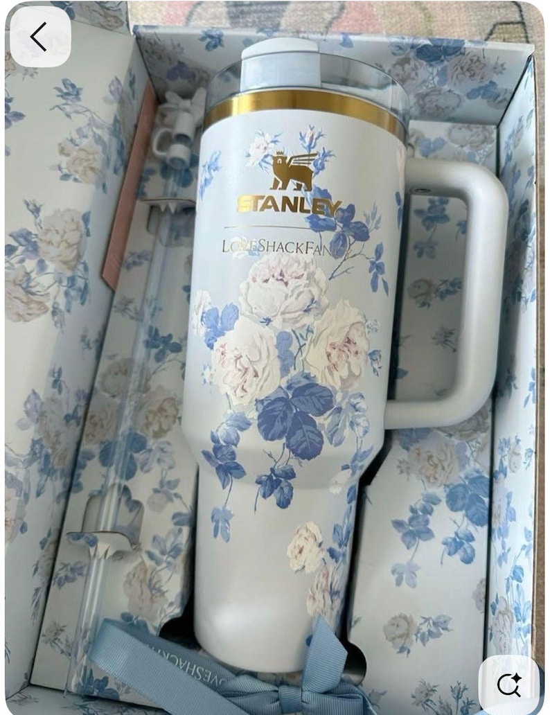 May include: A white Stanley tumbler with a gold rim and a blue and white floral pattern. The cup features the text Stanley and LoveShackFancy. It includes a matching clear straw with a white floral topper, all packaged in a floral box.