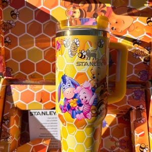 May include: A yellow Stanley tumbler featuring a honeycomb pattern and illustrations of Winnie the Pooh, Piglet, Eeyore, and Tigger. The cup includes a handle, a clear lid, and the Stanley logo printed in black on the front surface of the tumbler.