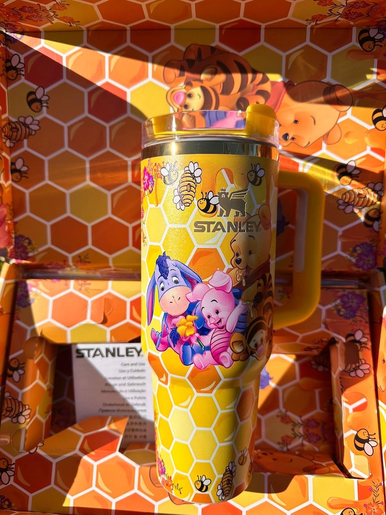 May include: A yellow Stanley tumbler featuring a honeycomb pattern and illustrations of Winnie the Pooh, Piglet, Eeyore, and Tigger. The cup includes a handle, a clear lid, and the Stanley logo printed in black on the front surface of the tumbler.