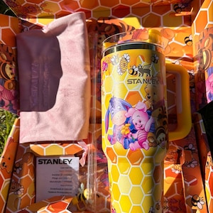 May include: A yellow Stanley tumbler featuring a honeycomb pattern and Winnie the Pooh characters, including Piglet and Eeyore. The set includes a clear straw, a light pink fabric pouch, and a Stanley care instruction card inside a themed box.