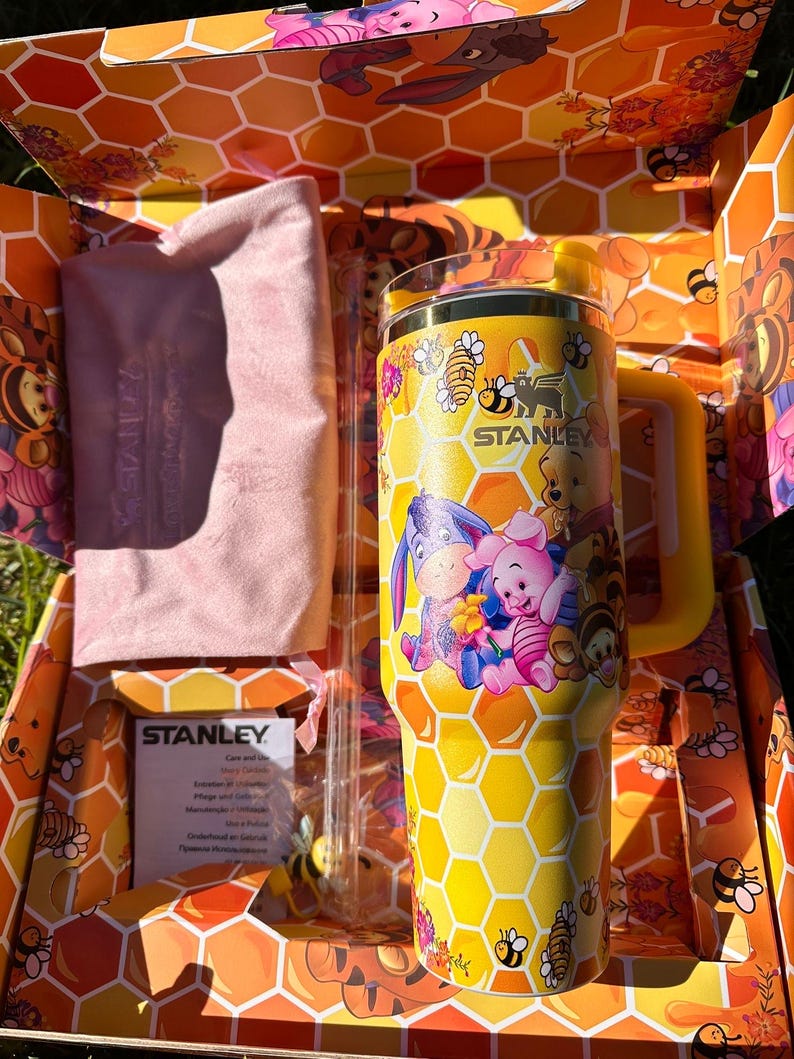 May include: A yellow Stanley tumbler featuring a honeycomb pattern and Winnie the Pooh characters, including Piglet and Eeyore. The set includes a clear straw, a light pink fabric pouch, and a Stanley care instruction card inside a themed box.