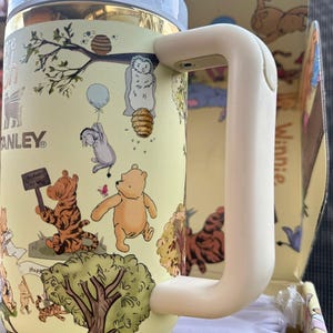 May include: A yellow insulated tumbler featuring Winnie the Pooh characters and scenes. The tumbler has a white handle and lid, with the "Stanley" logo visible. The design includes illustrations of Pooh, Tigger, and Eeyore, with bees and honeycombs.