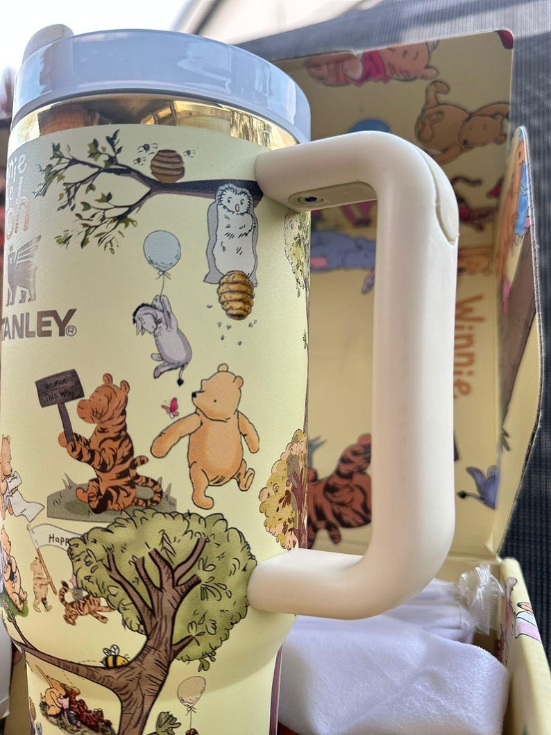 May include: A yellow insulated tumbler featuring Winnie the Pooh characters and scenes. The tumbler has a white handle and lid, with the "Stanley" logo visible. The design includes illustrations of Pooh, Tigger, and Eeyore, with bees and honeycombs.