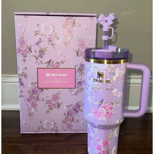 May include: A lavender Stanley tumbler with a floral pattern, a matching handle, and a straw topped with a bow. The tumbler features the Stanley and LoveShackFancy logos. It is displayed next to a matching rectangular box with the same floral design.