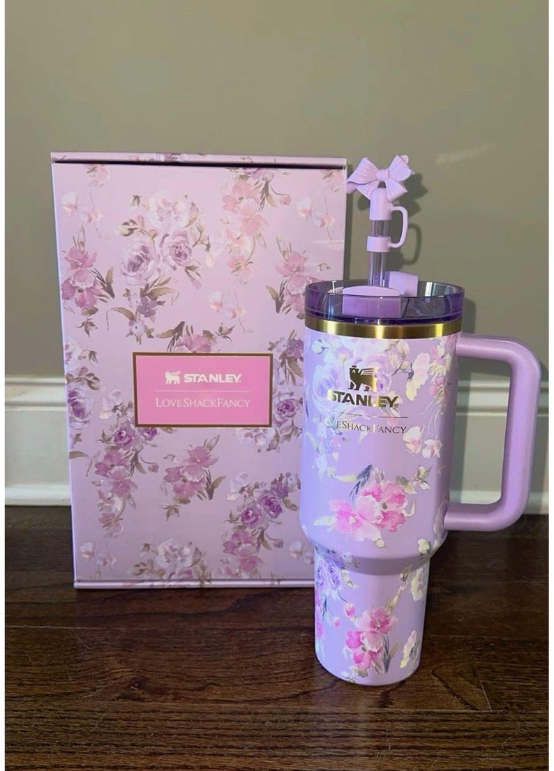 May include: A lavender Stanley tumbler with a floral pattern, a matching handle, and a straw topped with a bow. The tumbler features the Stanley and LoveShackFancy logos. It is displayed next to a matching rectangular box with the same floral design.